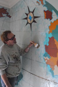 Faux artisan William Bell is applying imported Spatula Stuhhi beads to the world map mural giving the ocean a shimmering effect.